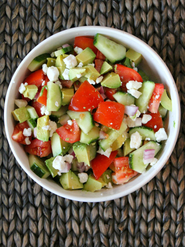 Overhead shot of avocado cucumber tomato salad in a white bowl placed on a dark brown woven place mat