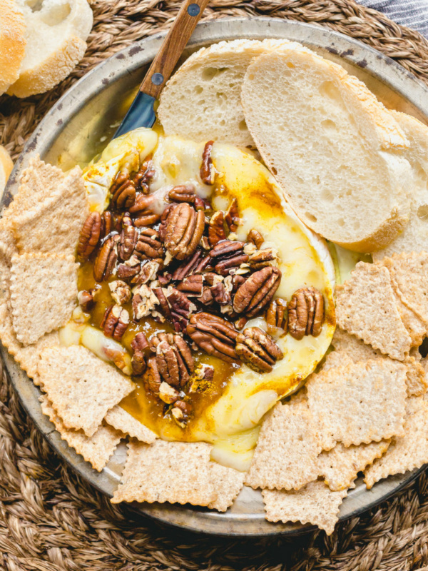 melted brie cheese with pecans on a platter surrounded by sliced baguette and crackers