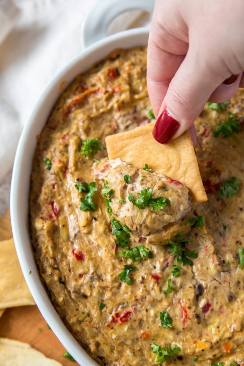 hand dipping cracker in white baking dish full of hot artichoke dip