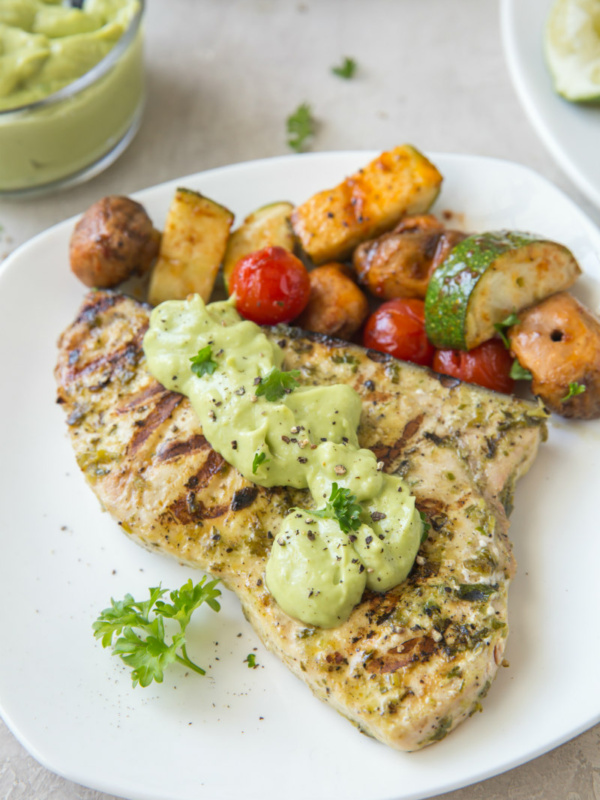 grilled swordfish topped with avocado mayonnaise on a white plate. Vegetables on the side.