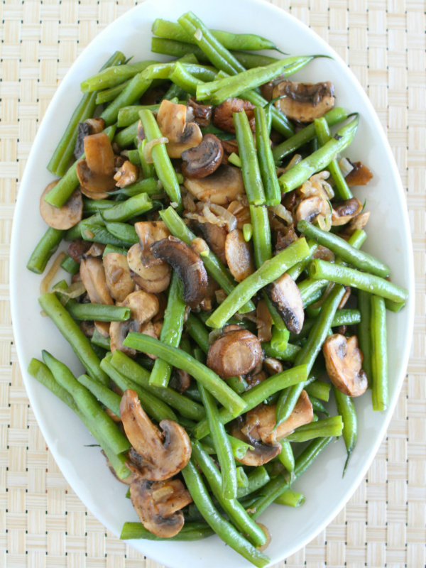 overhead shot of green beans with mushrooms and shallots on a white platter set on a woven white placemat