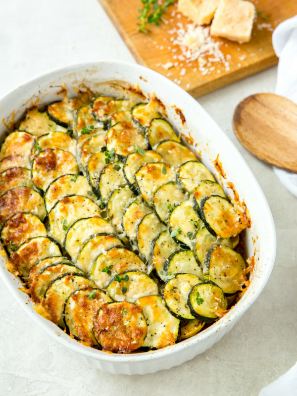 zucchini and tomato gratin in a white casserole dish with a wooden cutting board with cheese on it and a wooden spoon on the side