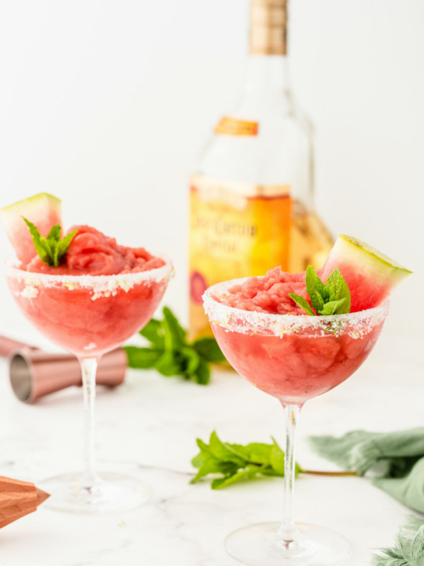 two watermelon mint margaritas garnished with mint and watermelon with a bottle of tequila in the background