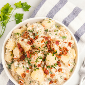 blue cheese and bacon potato salad in a white bowl with blue and white striped cloth napkin with parsley garnish