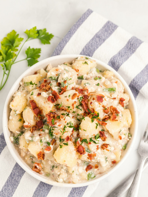 blue cheese and bacon potato salad in a white bowl with blue and white striped cloth napkin with parsley garnish