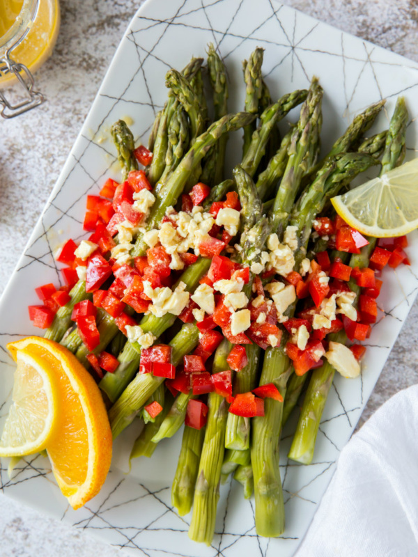 a white plate set on a marble background with chilled asparagus that are topped with feta vinaigrette with chopped red bell pepper and garnished with lemons and oranges