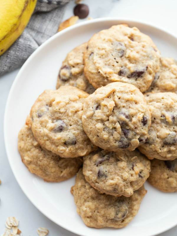 overhead shot of banana walnut cookies stacked on a white plate with a peek of a banana in the background