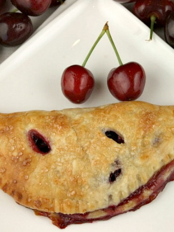 fresh cherry turnover on a white plate with a couple of fresh cherries on the plate. fresh cherries in the background