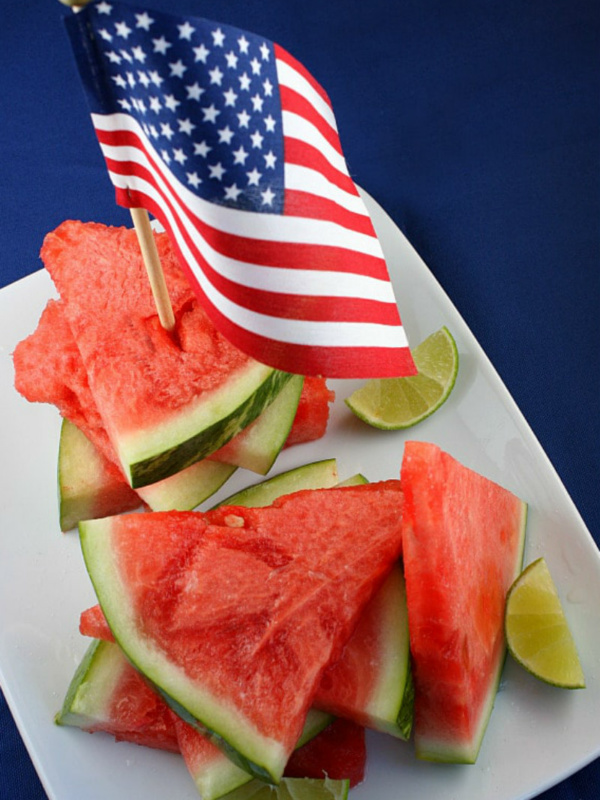 tequila soaked watermelon wedges on a white platter with lime wedges and an american flag stuck into the top