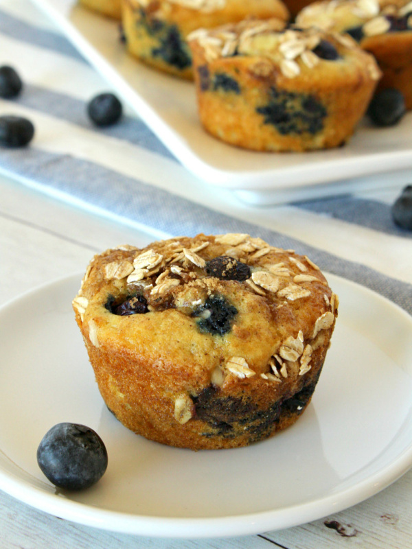 Blueberry Cinnamon Muffin on a white plate with more muffins on a platter in the background with a white and blue striped napkin