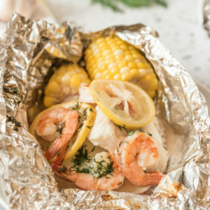 grilled new england seafood foil packets opened up to see the inside. fresh herbs in background