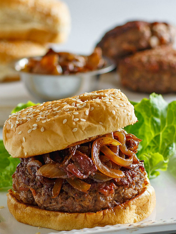 Bacon Burger with Balsamic Caramelized Onions on a white platter with a lettuce leaf. more buns, burgers and a bowl of caramelized onions in the background