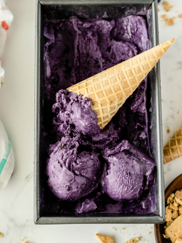 rectangular metal tub of blueberry cheesecake ice cream with a couple of scoops on top with an ice cream cone sitting in the tub