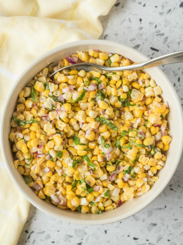 overhead shot of fresh corn salad in a white bowl with a serving spoon. light yellow napkin displayed on the side