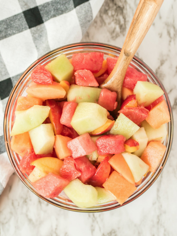 overhead shot of fruit salad in a glass bowl with a wooden spoon. black and white plaid napkin in background