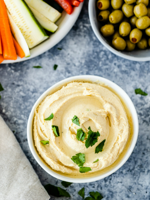 overhead shot of white bowls filled with hummus, fresh vegetables and olives