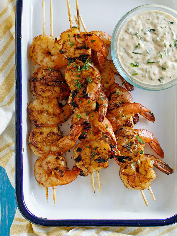 Barbecued shrimp on skewers with a bowl of remoulade sauce served on the side. Set on a white platter with a blue rim. Yellow striped napkin underneath