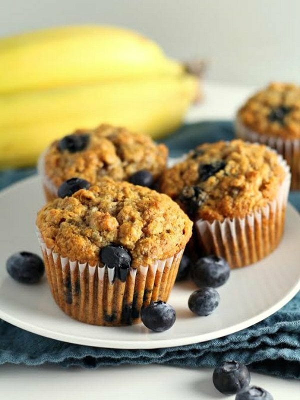 whole wheat banana blueberry muffins (3) on a white plate with fresh blueberries scattered about and bananas in the background