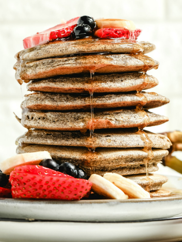 stack of buckwheat pancakes on a white plate with syrup drizzling down the sides, garnished with fresh berries and bananas