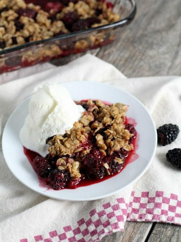 serving of blackberry crisp on a white plate with vanilla ice cream. Set on top of a white and red/white checked cloth napkin. Pan of crisp in the background + fresh blackberries