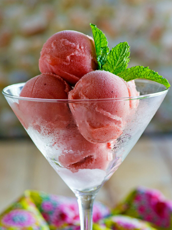 martini glass filled with three scoops of watermelon sorbet garnished with fresh mint leaf sprig. colorful pink and green patterned cloth napkin displayed underneath.