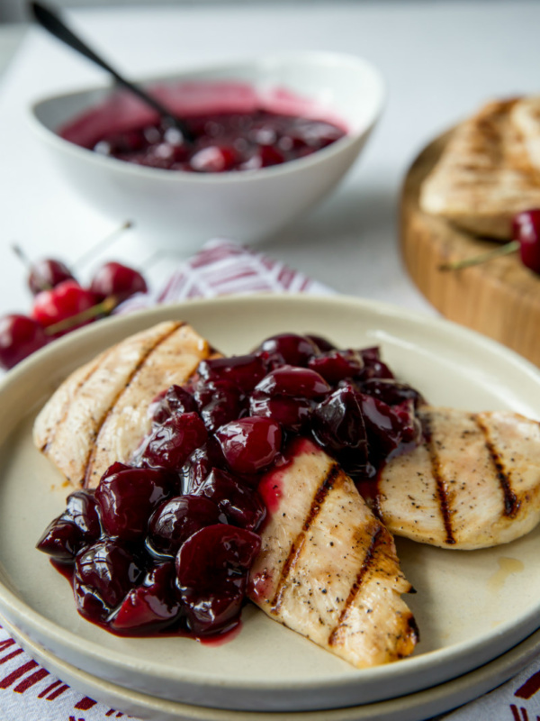 grilled chicken on a white plate with cherry sauce . bowl of cherry sauce in the background