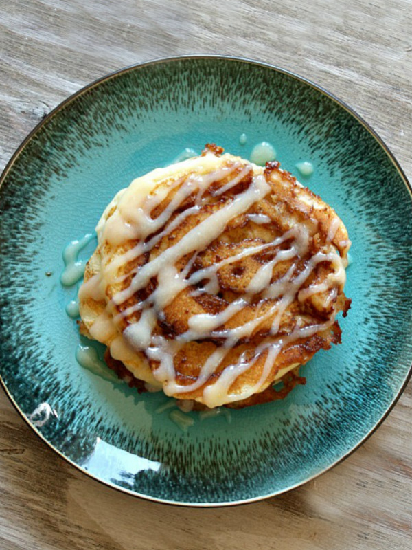 overhead shot of cinnamon roll pancakes with cream cheese drizzle on a teal green plate on a wooden board