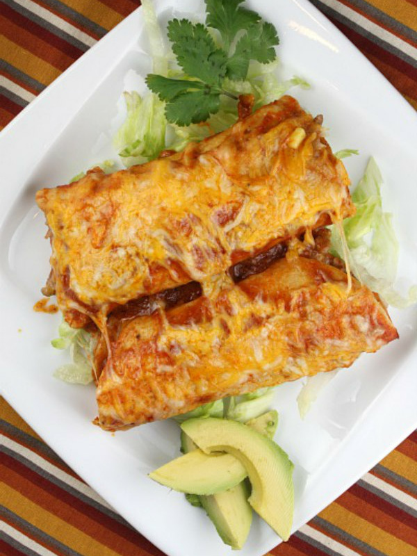 two beef enchiladas on a white plate with avocado and cilantro garnish- set on a striped placemat