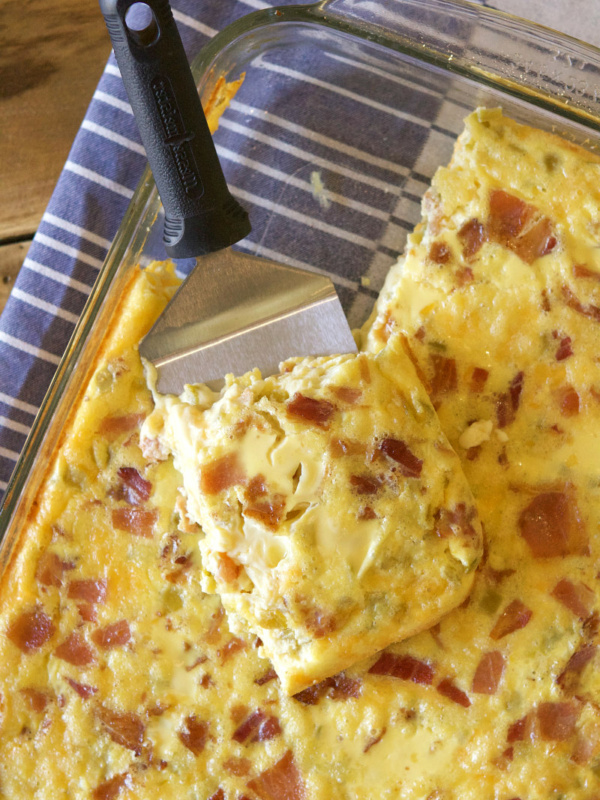 green chile bacon and cheese egg bake in a pyrex casserole dish with a spatula serving it. Sitting on a blue and white striped cloth napkin