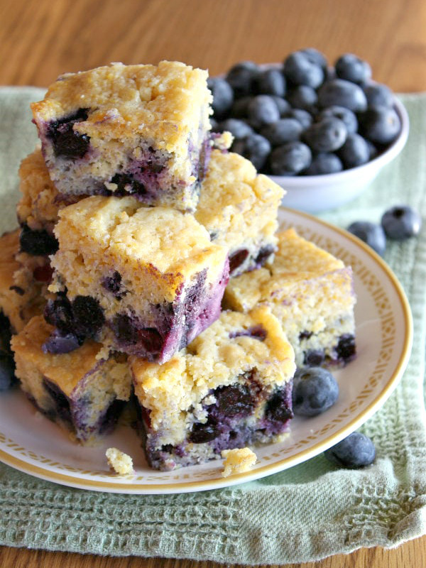 pieces of blueberry cornbread stacked on a plate with a bowl of blueberries in the background- set on a green towel on a wooden surface