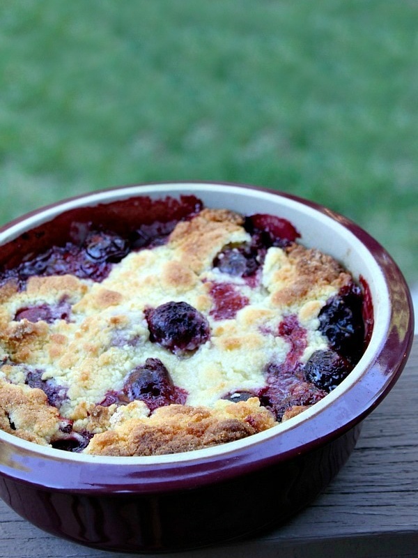 cherry cobbler in a round burgundy casserole dish sitting on a wooden railing with green background