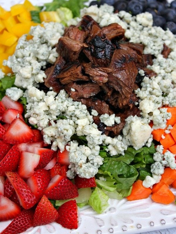 grilled steak summer salad on a white plate with steak piled in the middle surrounded by blue cheese and vegetables and fruit