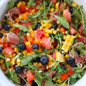 overhead shot of blueberry corn salad with prosciutto in a white bowl on a green background