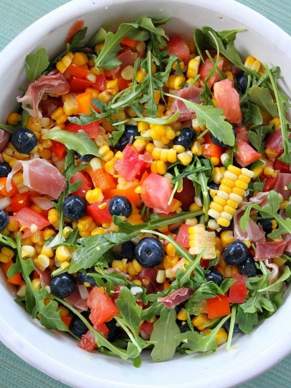 overhead shot of blueberry corn salad with prosciutto in a white bowl on a green background