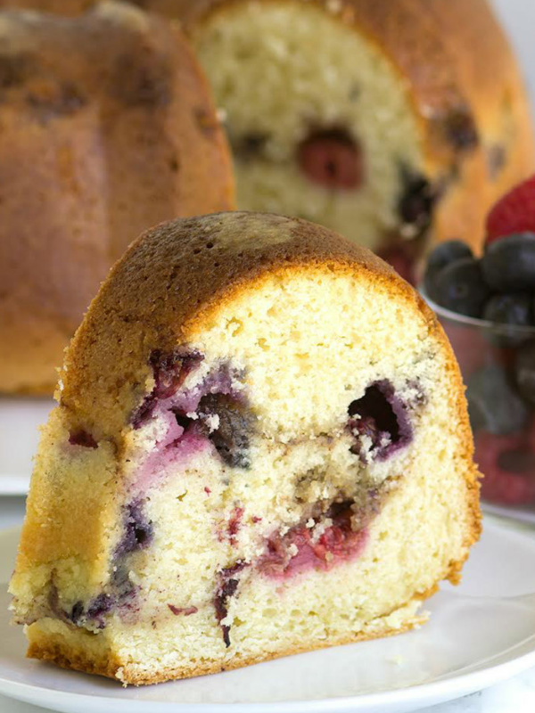 slice of two berry coffee cake on a white plate in front of the rest of the coffee cake