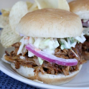 slow cooker pulled pork sandwich on a white plate, garnished with red onion and cole slaw with potato chips on the plate. Blue napkin underneath