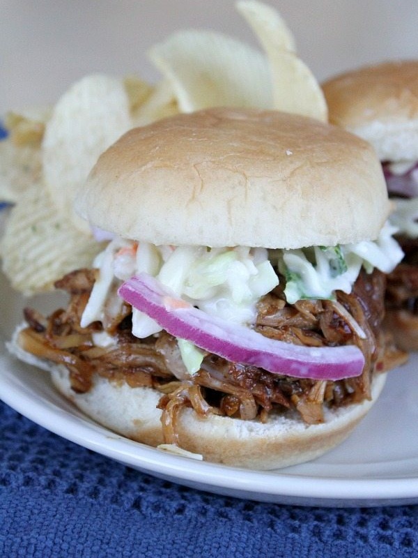 slow cooker pulled pork sandwich on a white plate, garnished with red onion and cole slaw with potato chips on the plate. Blue napkin underneath