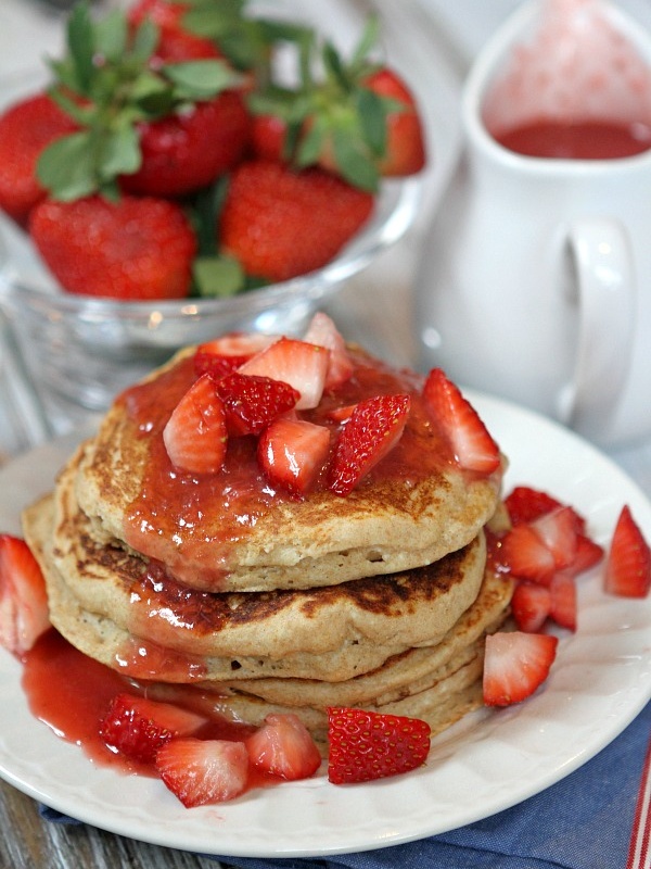 stack of strawberry ricotta pancakes. lots of fresh strawberries and strawberry sauce on top with fresh strawberries and pitcher of sauce in background.