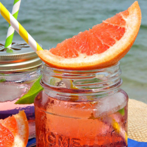 ruby sunset cocktail in a small glass jar garnished with fresh grapefruit, another cocktail next to it. Sitting on a blue napkin with white polka dots with the ocean in the background