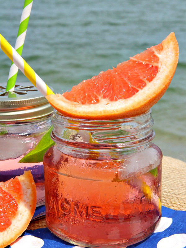 ruby sunset cocktail in a small glass jar garnished with fresh grapefruit, another cocktail next to it. Sitting on a blue napkin with white polka dots with the ocean in the background