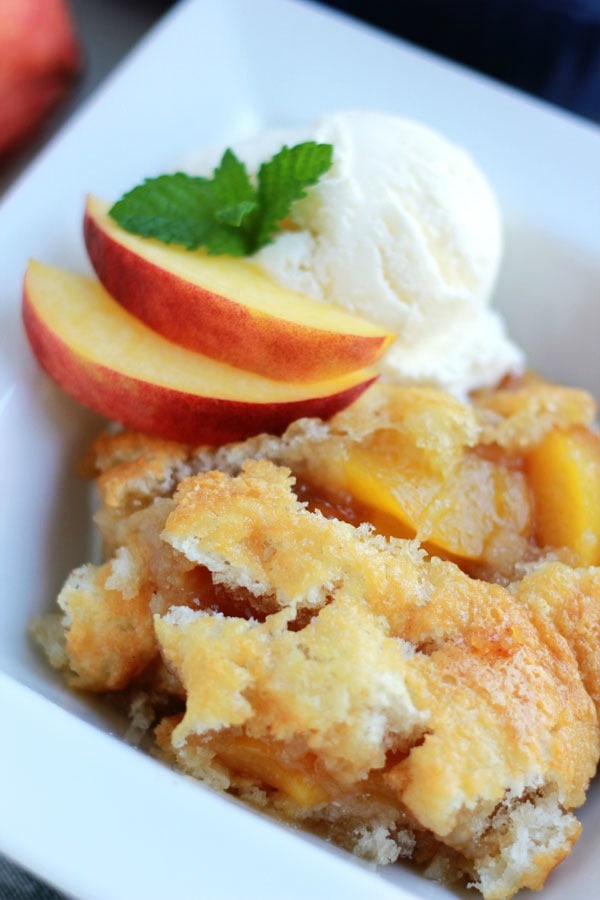 overhead shot of a serving of peach cobbler in a white serving dish garnished with ice cream, fresh peaches and mint leaves