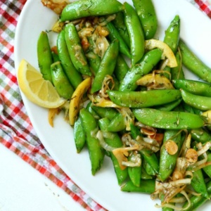 overhead shot of lemony stir fried sugar snap peas on a white plate garnished with a lemon wedge. Set on a multi-colored checked cloth napkin