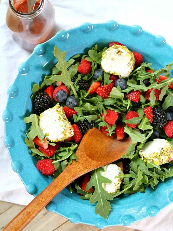 overhead shot of arugula berry and goat cheese salad in a blue bowl with a wooden serving spoon and a bottle of dressing on the side