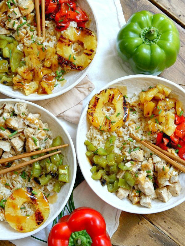 overhead shot of 3 white bowls filled with grilled chicken and pineapple rice bowls with chopsticks. Set on a white napkin with a wood board background with fresh bell peppers and green onions garnish.