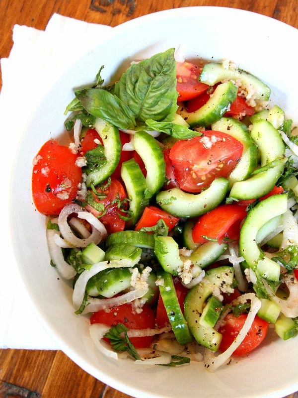 Overhead shot of Tomato Cucumber and Basil Salad in a white bowl with a white napkin on a wood surface