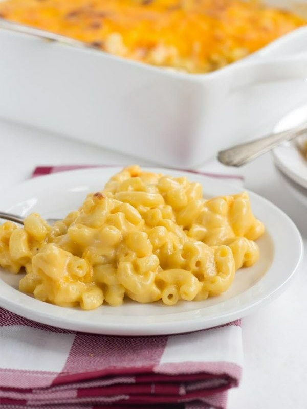 macaroni and cheese on a white plate set on a red and white plaid napkin with white casserole dish of macaroni and cheese in the background