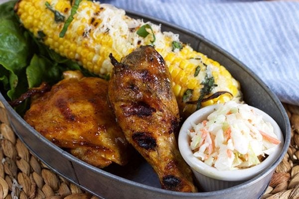 carolina style barbecue chicken in a serving dish with cole slaw and corn on the cob on a woven mat with a blue/white striped napkin