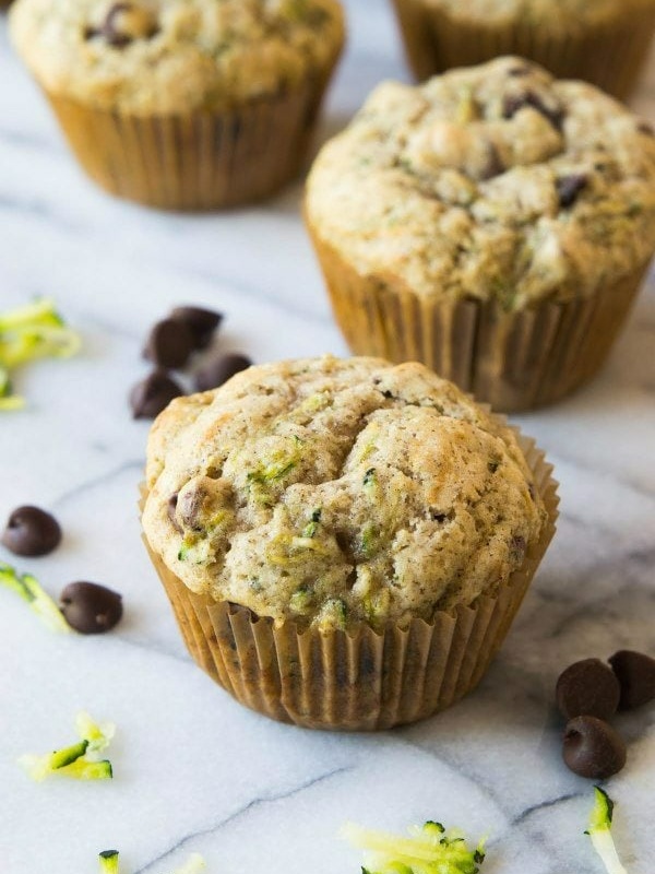 zucchini chocolate chip muffins sitting on a marble surface with chocolate chips and shredded zucchini scattered about