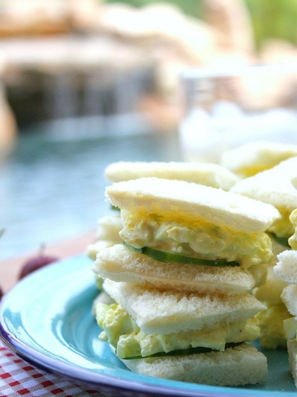 avocado egg salad tea sandwiches stacked on a blue plate on a red/white checked tablecloth with pool backyard setting