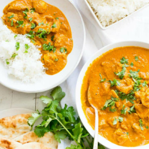 overhead shot of two servings of indian butter chicken in white bowls with white rice and cilantro garnish and pita bread on the side